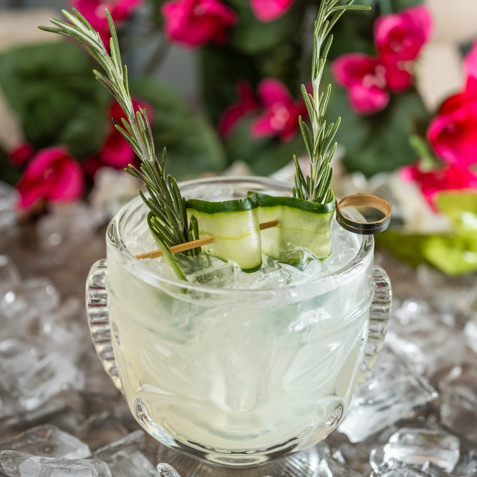 Icy pale cocktail garnished with cucumber ribbons and rosemary sprigs, surrounded by ice and pink flowers.