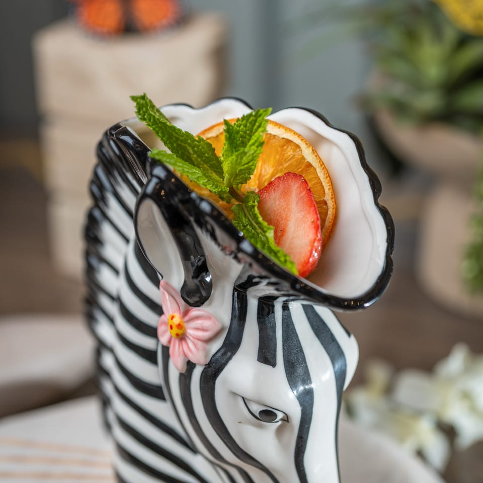 Zebra-shaped dessert cup filled with cream, orange slices, strawberries, and mint, with a butterfly and plants in the background.