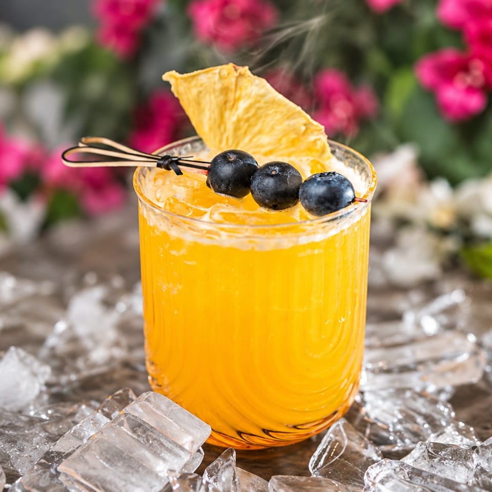 Bright orange cocktail garnished with blueberries and a citrus chip, chilling among ice against a colorful floral backdrop.