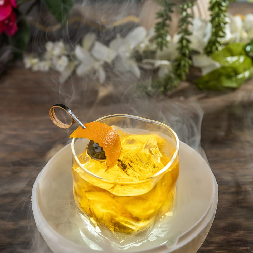 Golden cocktail with ice, orange peel garnish, and swirling smoke, served in a glass on a white plate amid tropical greenery.