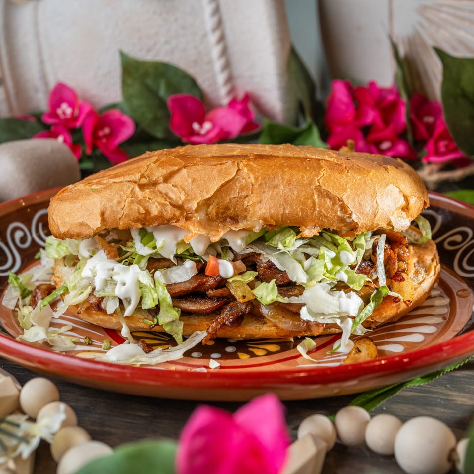 Loaded grilled meat sandwich with lettuce, tomato, onions, and creamy sauce on a decorative plate surrounded by flowers.