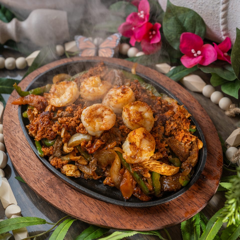 Steaming skillet of grilled shrimp, ground meat, peppers, onions, and vegetables on a wooden board surrounded by flowers and greenery.