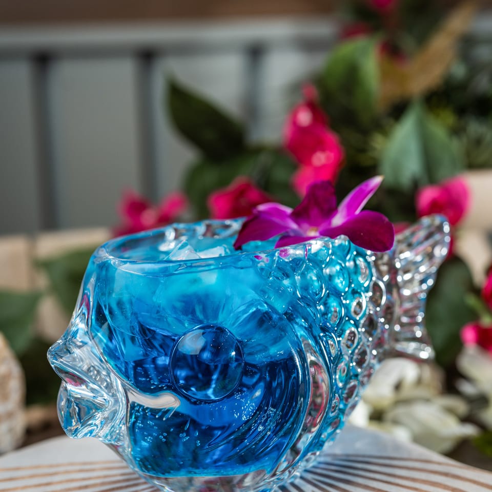 Blue cocktail in an ornate glass garnished with purple orchids, set on a patterned table beside tropical plants.