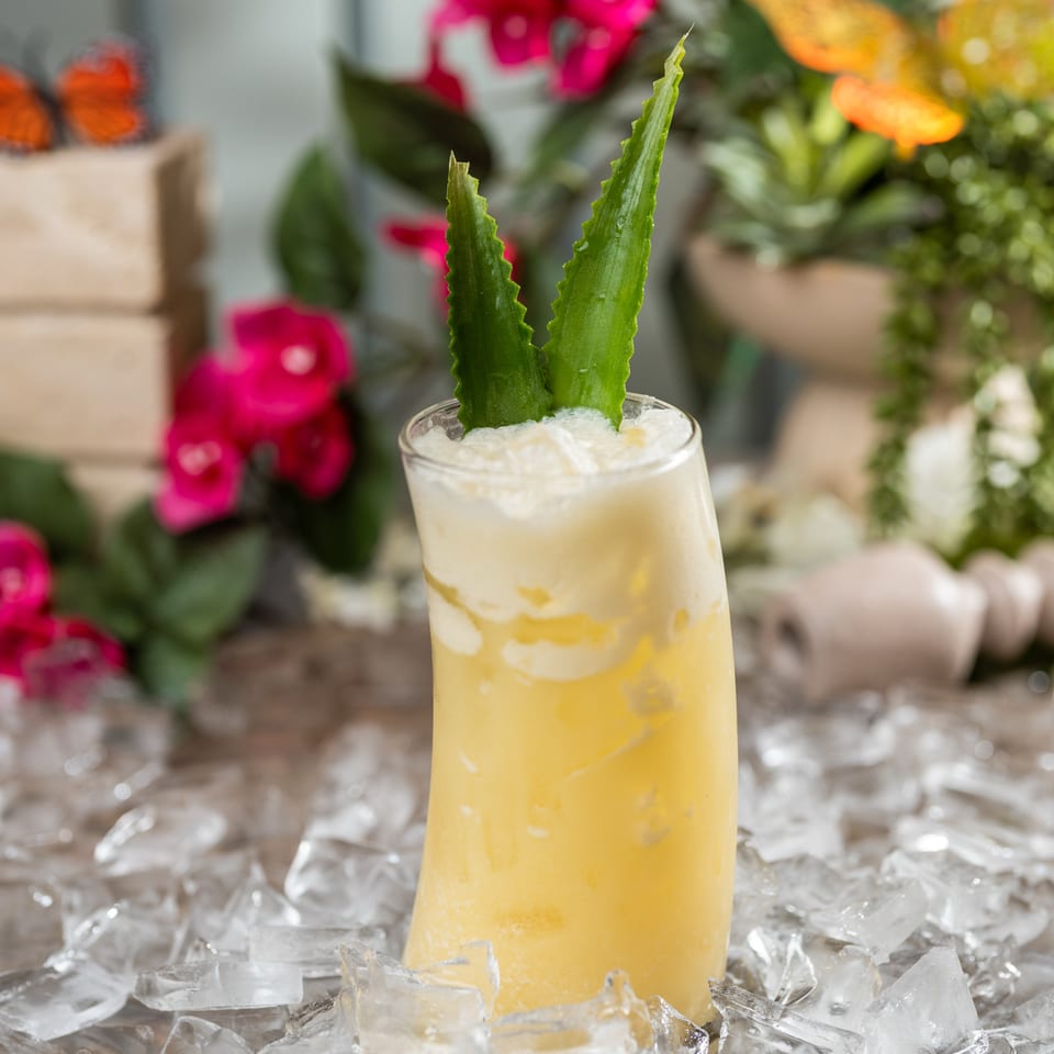 Yellow frothy drink garnished with aloe leaves, displayed in a glass surrounded by ice and colorful flowers.